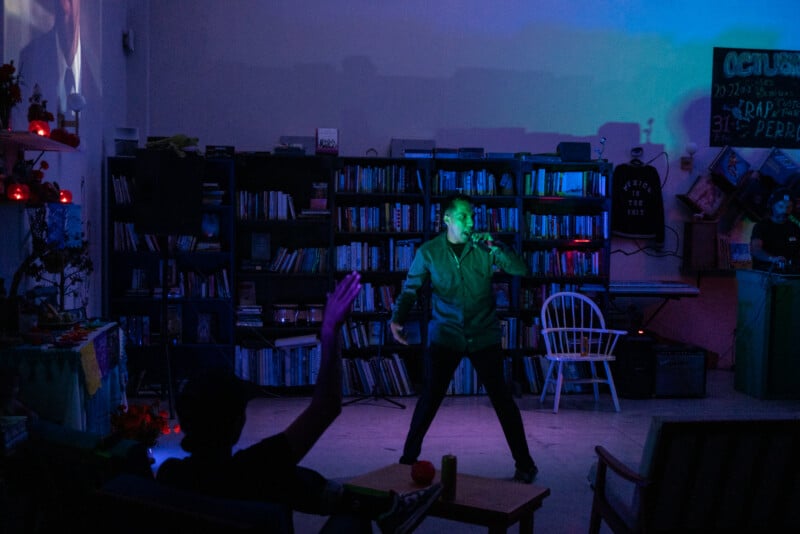 A person performs on a small stage in front of bookshelves, holding a microphone under blue and green lighting. An audience member raises a hand from a seated area in the foreground.