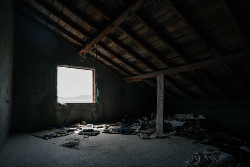A dimly lit, abandoned attic with a slanted wooden roof, debris, and scattered clothes on the floor. Light shines through a single open window, illuminating the neglected space.