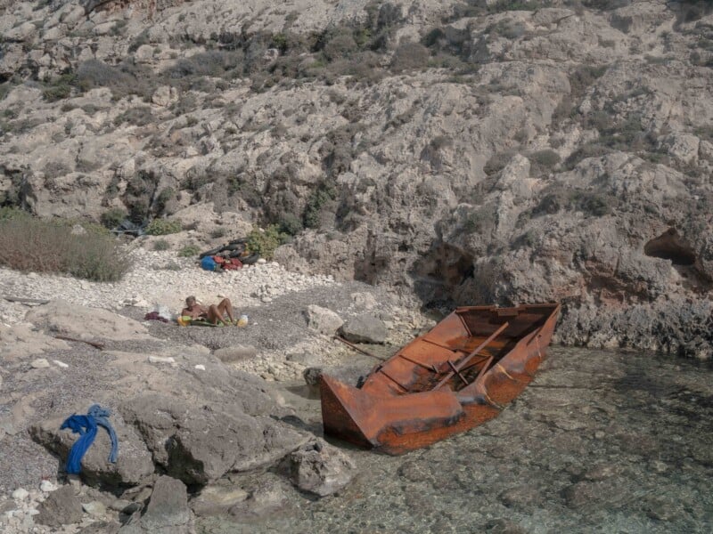 A rusted, abandoned boat rests in shallow, clear water near rocky terrain. A person lies on a towel sunbathing on the pebbled shore, with clothes and belongings scattered nearby.