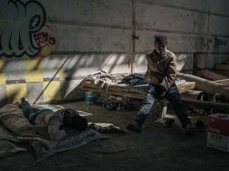 Two people in worn clothes rest inside an abandoned, dimly lit building. One person lies on a blanket on the floor, while the other sits on a broken bed. Sunlight streams in, highlighting the graffiti-covered wall behind them.