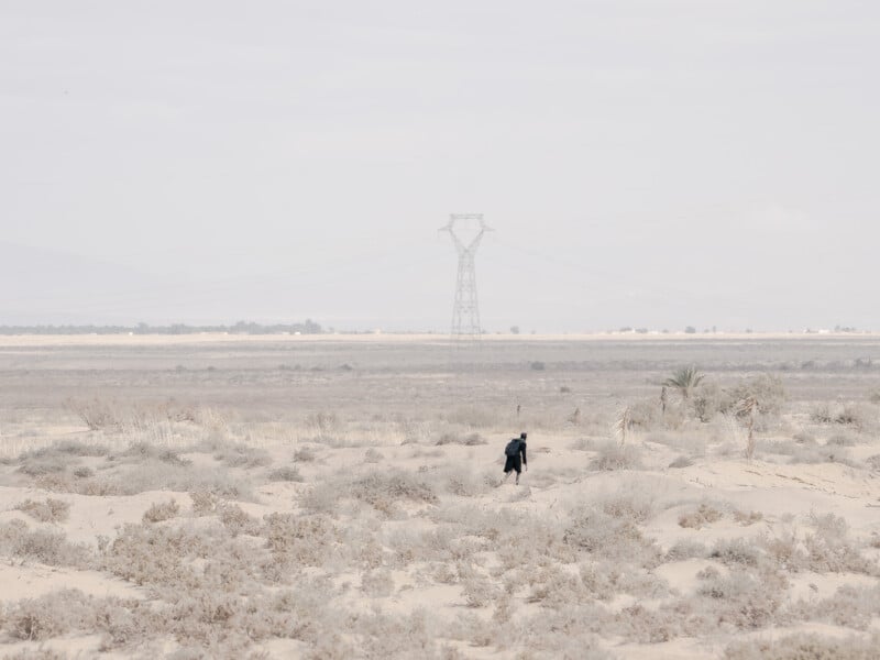 A person dressed in black walks alone through a dry, sandy landscape with sparse vegetation; a large powerline tower stands in the distance under a pale, hazy sky.