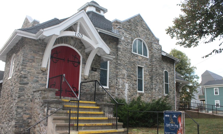 An old stone building that was previously a church with concrete steps bordered by a wrought iron railing leading up to its dark red door.