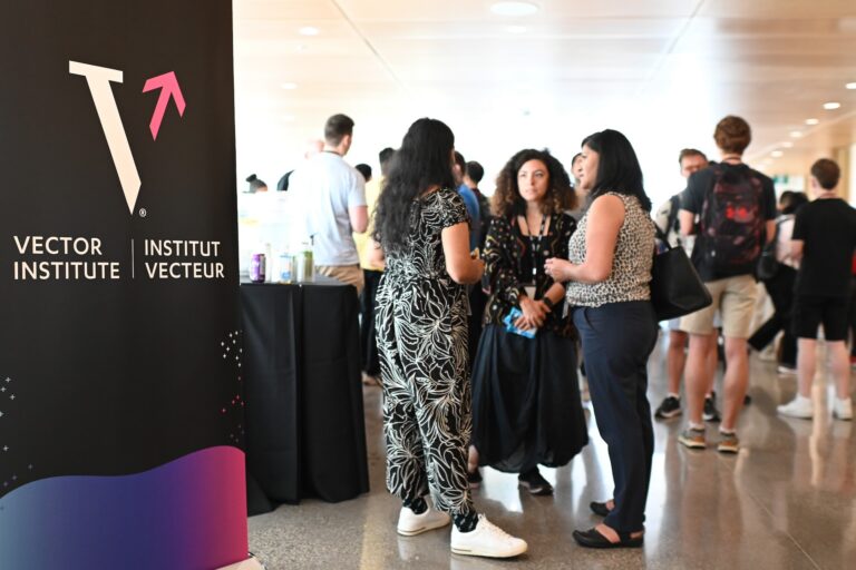 Three people facing each other while standing and talking. There are blurred-out people in the background and a sign on the left displays the Vector Institute logo.