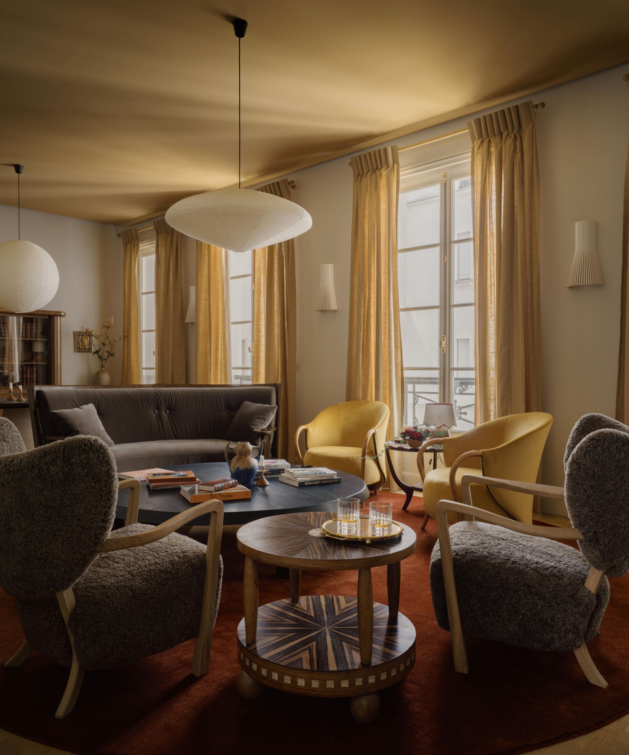 A living room with an ochre ceiling, warm white walls and large window with ochre curtains. A dark red rug and dark neutral chairs.