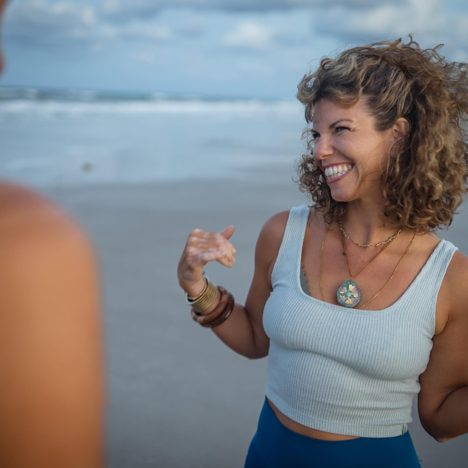 Woman in tank top smiling at someone with beach and sea in background
