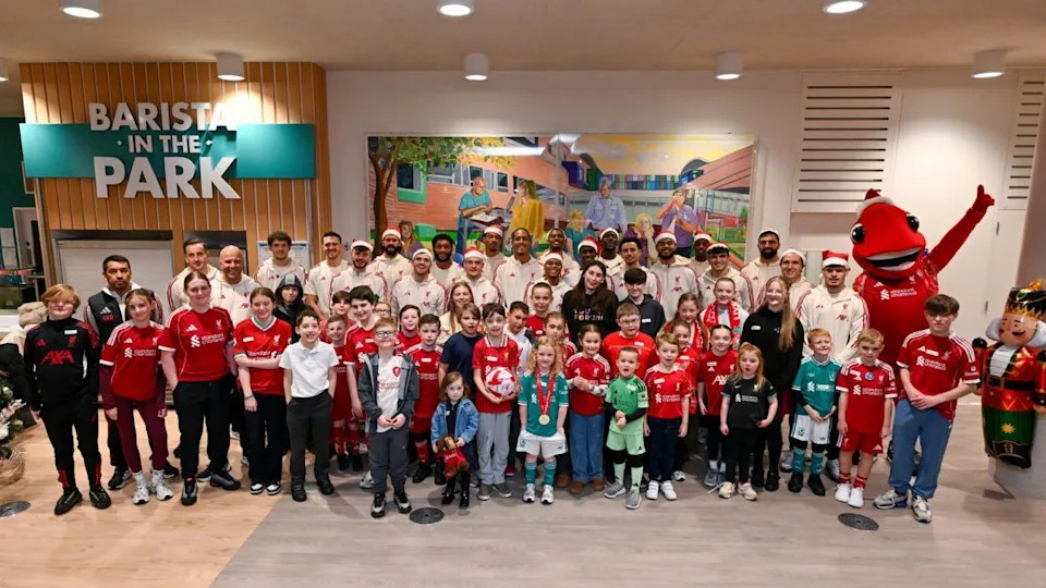 Patients wearing Liverpool kits are smiling as they stand in front of the smiling Liverpool squad members in a large room, with a Christmas tree and a large nutcracker figurine at either side of them.
