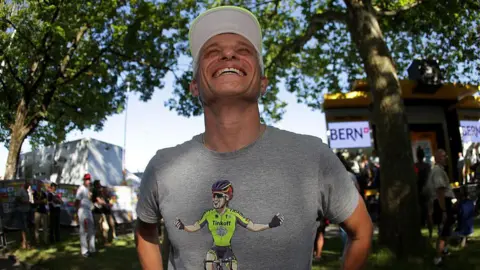 Chris Graythen/Getty Images A man wearing a white baseball cap and grey T-shirt grins while standing under a tree