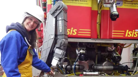 Supplied Dr Taylor wearing a white hard hat, blue jumper and yellow apron. She is standing behind a large piece of red and yellow machinery.