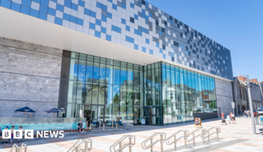 The exterior of The Box museum and art gallery. The ground floor has large glass panel windows. There are people sat outside of the museum and people walking to the right. There is a blue square design above the windows.