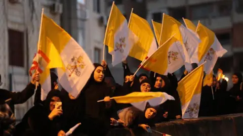 Getty Images Excited young women in Islamic scarves wave papal flags outside a building.
