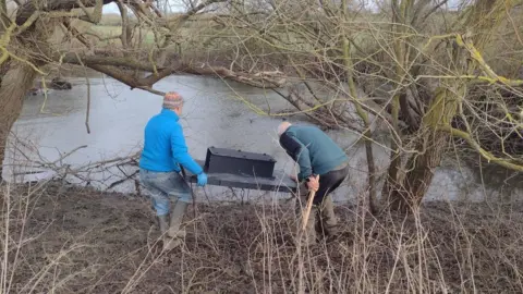 RTCT Two volunteers deploying a raft on the river. They have their backs to the camera. It's an overcast day.