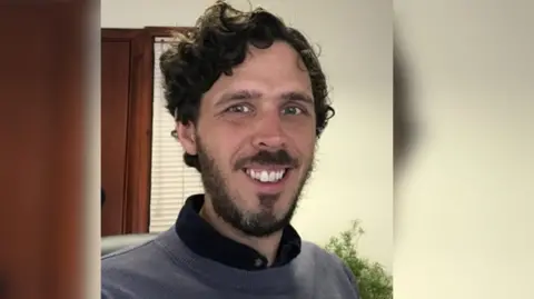 Nosa A selfie of James Hicks wearing a grey sweatshirt with a black collar poking out the top. He has green-grey eyes, black curly hair and a trimmed beard, and is smiling at the camera. Behind him there is a cream wall, a wooden doorway and a plant.