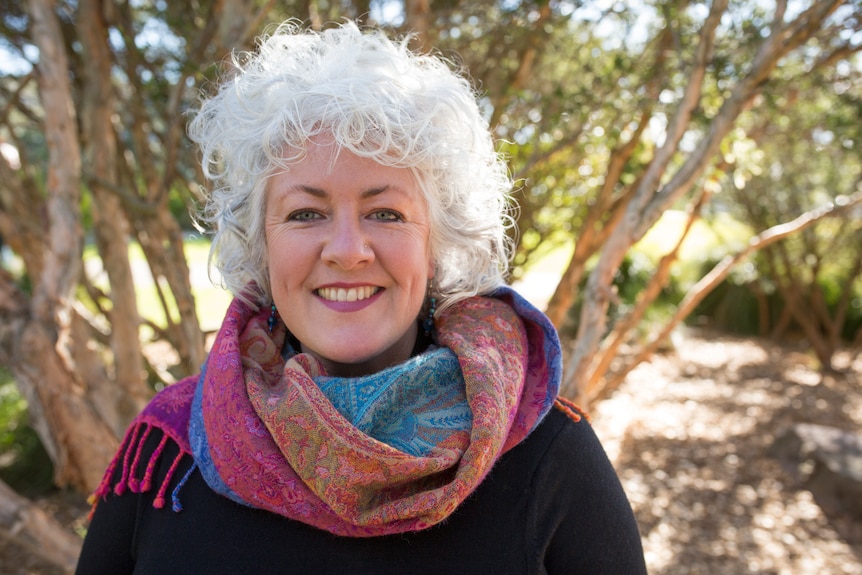 A woman with white curly hair smiles with a tree in the background.