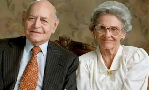 An elderly man in a suit and tie sits next to an elderly woman in a blouse and glasses, both smiling at the camera in a formal setting