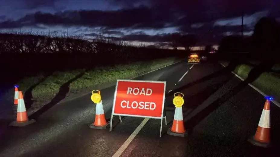 A straight section of country road pictured at night. In the foreground are cones and a sign stating, 'Road Closed'. Emergency vehicles are in the distance.