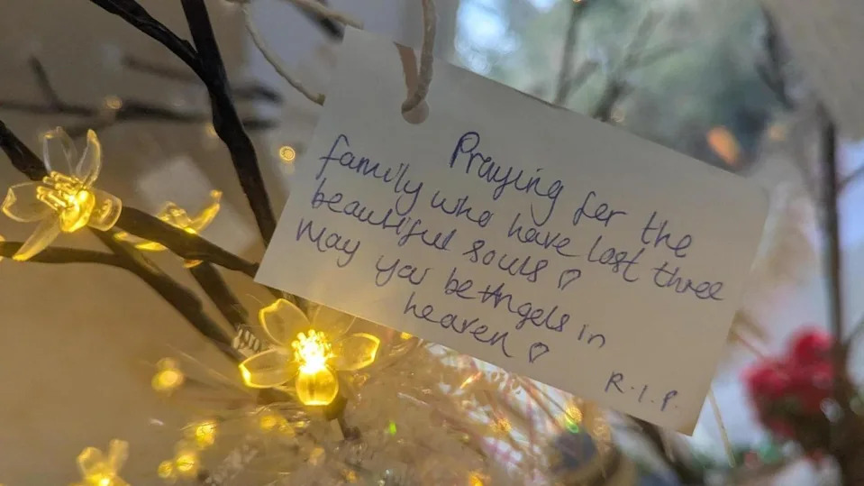 Close up of tributes to the family, which reads "praying for the family who have lost three beautiful souls. May you be angels in heaven." The card is attached to branches with a lights around.