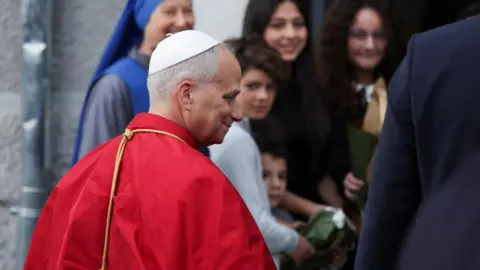 Reuters A man in a red mantle and white skullcap smiles outside a building where a small crowd of excited women and children stand.   