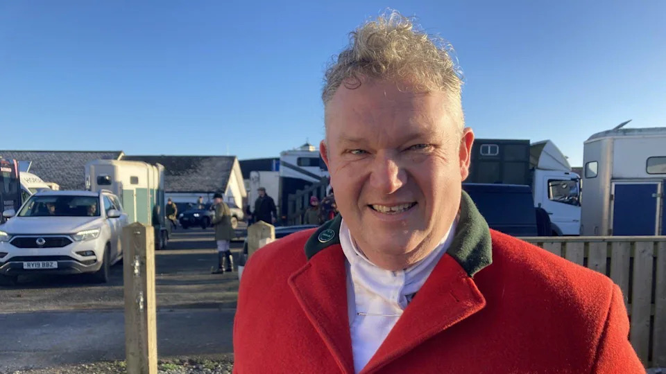 Gerrard Farmer stood smiling at the camera wearing a red coat and a white shirt. He has short light brown hair. People on horses can be seen behind him. The sky is blue. 
