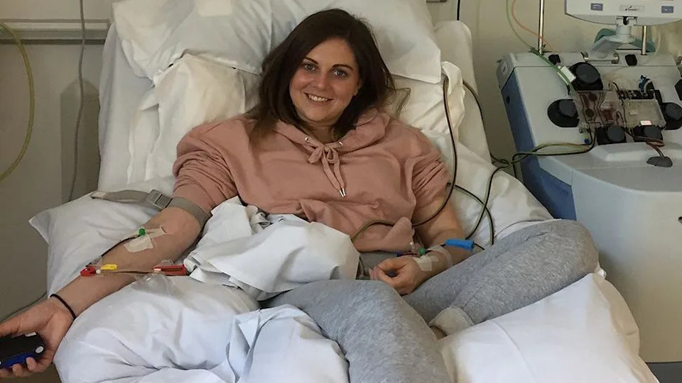 Holly Murdey, lying but with her back propped up, on a hospital bed. She's wearing a pastel pink hoodie and grey jogging bottoms, has dark shoulder-length hair and is smiling. Her arms have intravenous tubes attached with tape, with yellow, red and green valves. To her left is a piece of hospital equipment, into which some of the tubes are fed.
