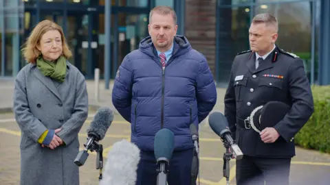PA Chloe Turner, Leader of Stroud District Council, Det Supt Ian Fletcher and  deputy chief fire officer Nathaniel Hooton. They are standing in front of five microphones outside the police headquarters.