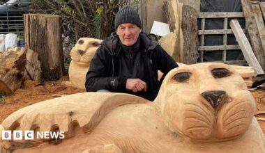 Ste Iredale is crouching behind one of the new seal sculptures. It has been hand-carved and there is a second one behind Mr him. He is wearing a hat and a black coat.