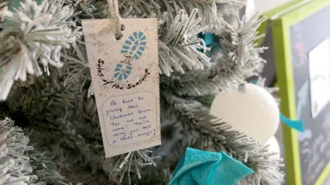 A message is pinned to an artificial, Christmas tree, alongside the more traditional baubles.  It tells people "Be kind to yourself - you are not alone."