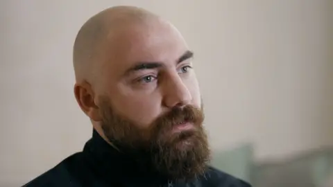 Konstantine has a shaved head, a reddish beard and blue eyes and is wearing a dark top