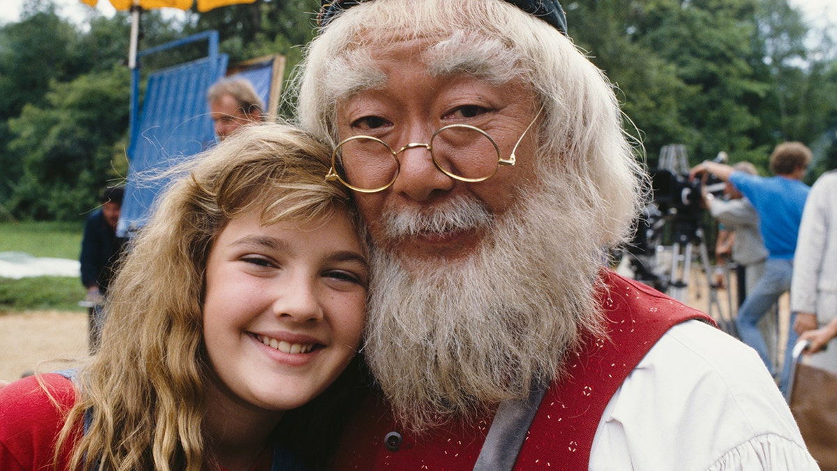 A smiling Drew Barrymore being hugged by Pat Morita.