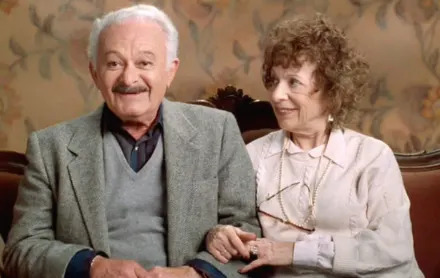 Elderly man and woman sitting on a couch, holding hands and smiling, with floral wallpaper in the background