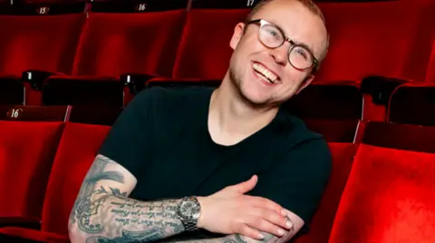 Danny Kaan Joe in a black top, wearing glasses, sitting in red theatre/cinema seats