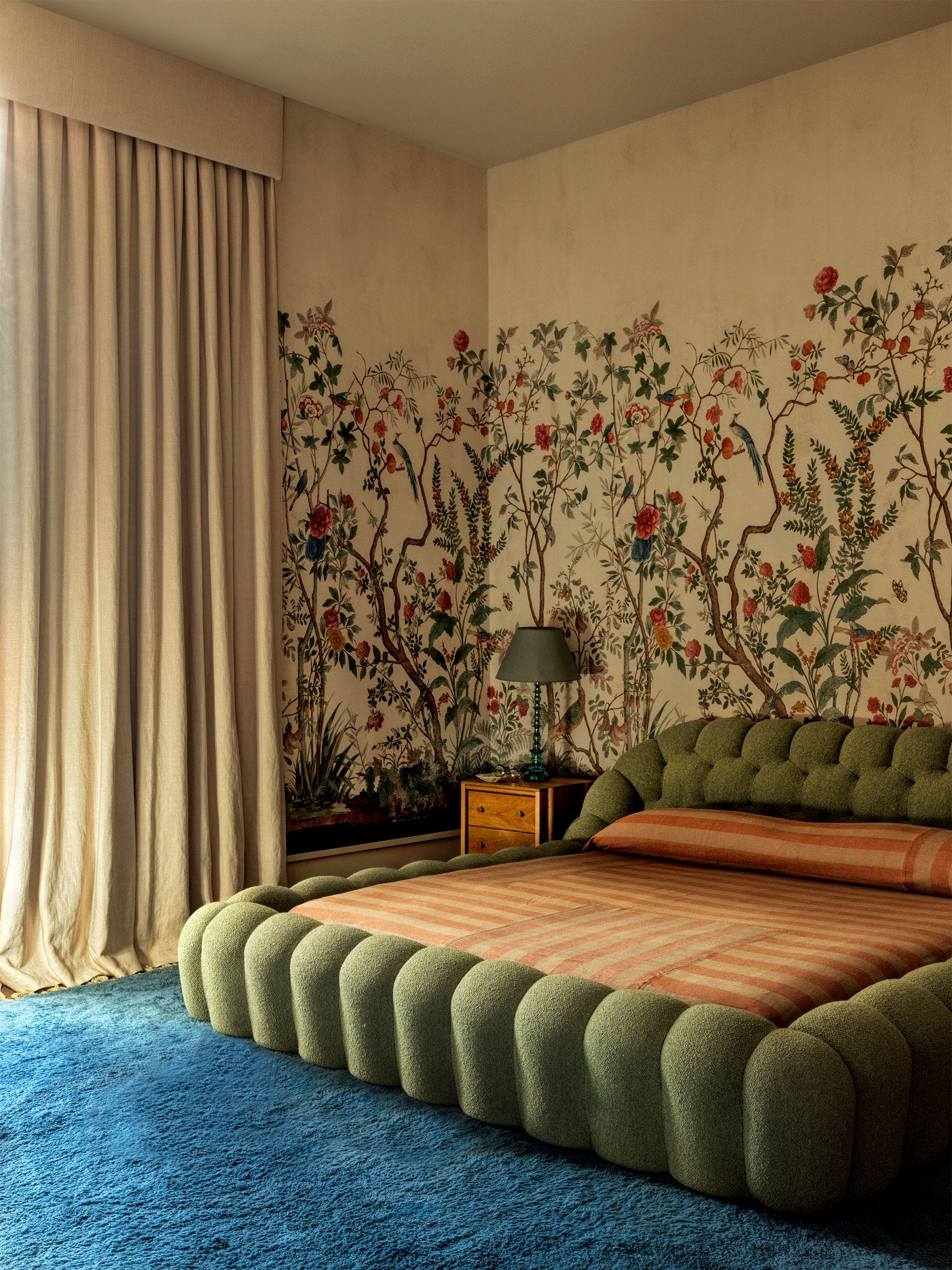 bedroom with a blue carpet, tufted olive green bed with striped orange and tan bedding, tan drapes that match the background wallpaper color, wallpaper in a viney, flowery print, nightstand with olive green lampshade
