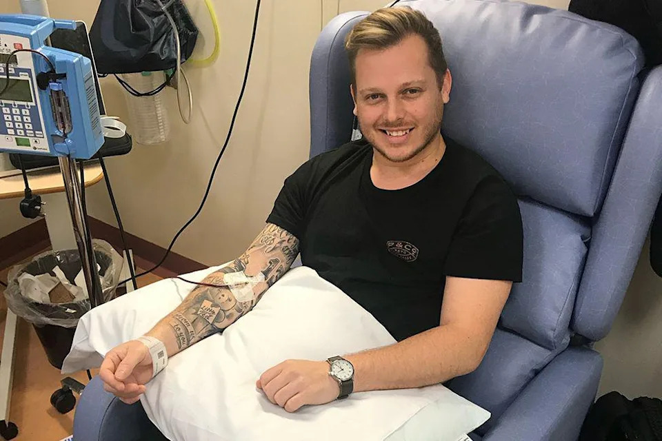 Ben Buttley, sitting in a cornflower-blue hospital reclining chair and smiling. He is wearing a black t-shirt and has short dark blonde hair, swept back at the front, with a short moustache and beard. He has a black-strapped analogue watch on his left wrist and his right arm - which is covered in tattoos - is resting on a white pillow on his lap. A tube is fed into it and is taped in place. He has a hospital identity bracelet on his right wrist. A piece of hospital equipment, on a metal stand, is on his right.