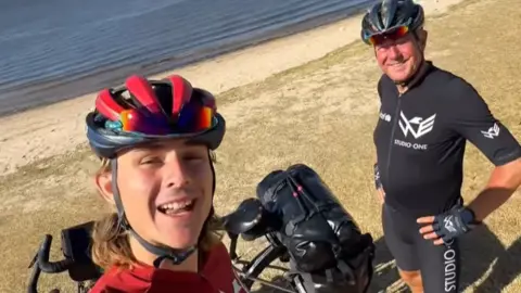 Josh Kohler/Instagram A selfie image of two men in cycling gear, one in red and one in black. They are standing by a beach