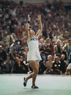 Young woman raises her hands in celebration on a tennis court before a large crowd.