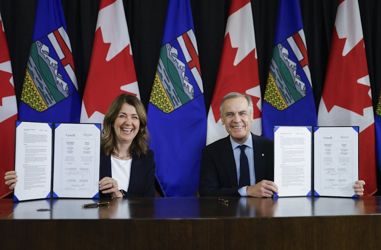 A man and woman wearing business attire sit at a table displaying documents. Behind them are flags of canada and alberta on poles.