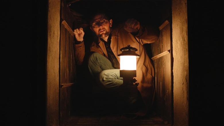 Adam Scott as Ohm Bauman crouching and holding a lantern in Hokum