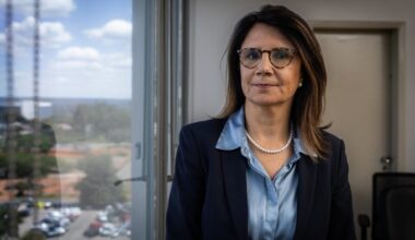 Ana Melia Campos Toni stands indoors near a window, wearing glasses, a navy blazer, a blue blouse, and a pearl necklace.