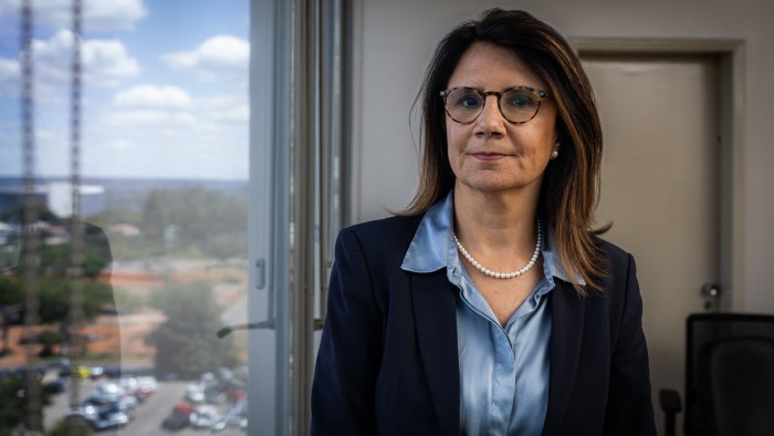 Ana Melia Campos Toni stands indoors near a window, wearing glasses, a navy blazer, a blue blouse, and a pearl necklace.