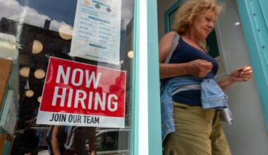 A "Now Hiring" sign is displayed in a business window as a woman exits the storefront.