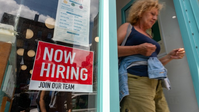 A "Now Hiring" sign is displayed in a business window as a woman exits the storefront.