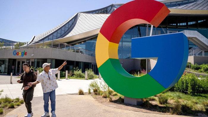 Two people walk past a large Google logo sculpture in front of the Bay View campus building in Mountain View, California.