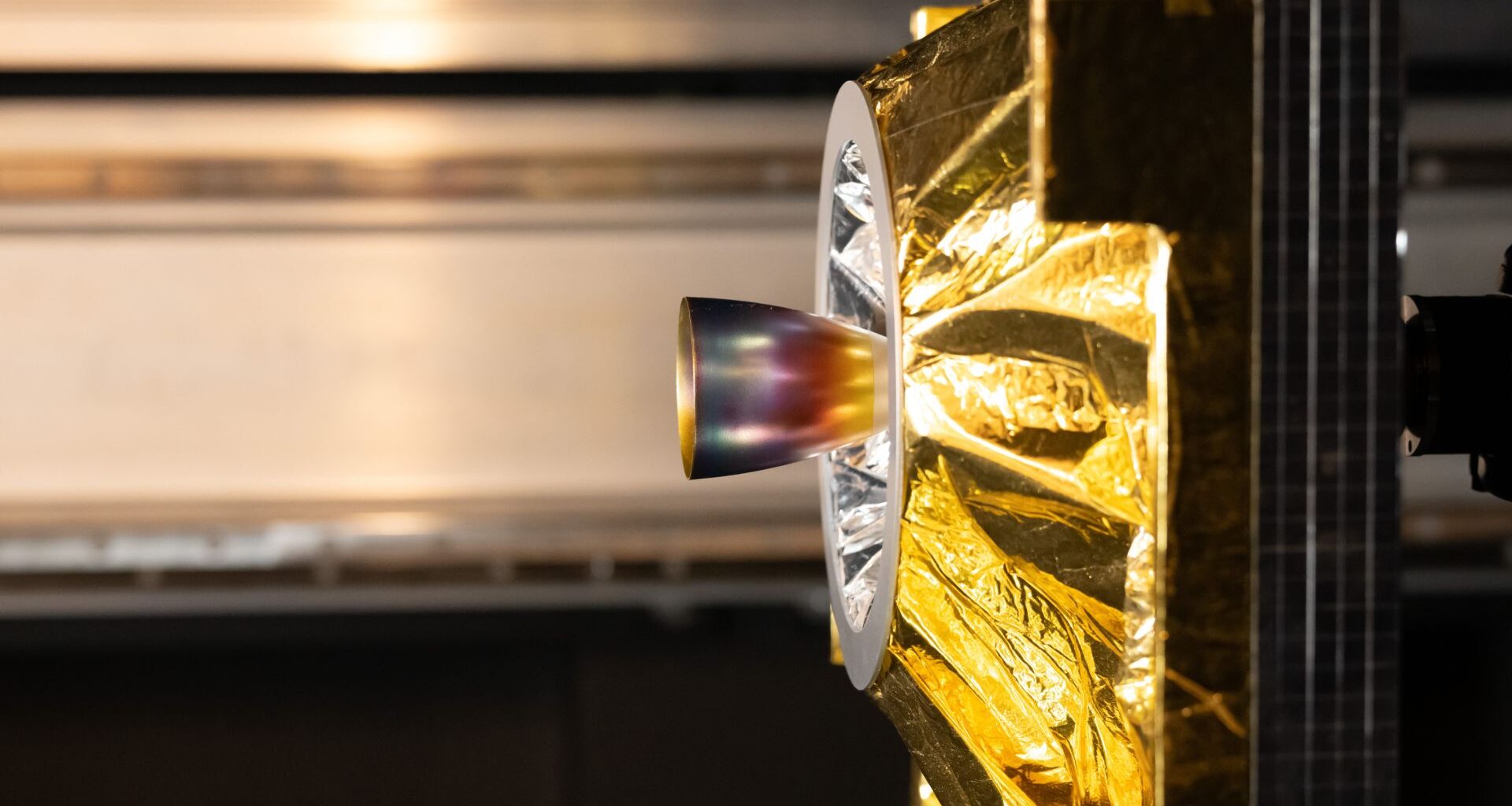 This is a close-up photo of a satellite model, seen from the side. A prominent feature is a cone-shaped nozzle extending from the centre of the model. The nozzle has an iridescent surface with shades of blue, purple, red and gold. The rest of the model is covered in crinkled gold-coloured thermal insulation, which is shiny and reflective. The background is out of focus, but horizontal metal rails mounted on the wall behind the model are visible. The lighting is bright and highlights the textures of the nozzle and the insulation.