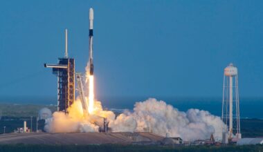 a white and black rocket launches into a clear blue sky from its ocean side launch pad