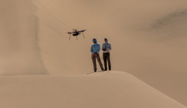Two people stand atop a sand dune, facing the camera. They are both looking at a drone flying in front of them. The person on the right holds a controller. Other sand dunes behind them make up the background of the entire image.