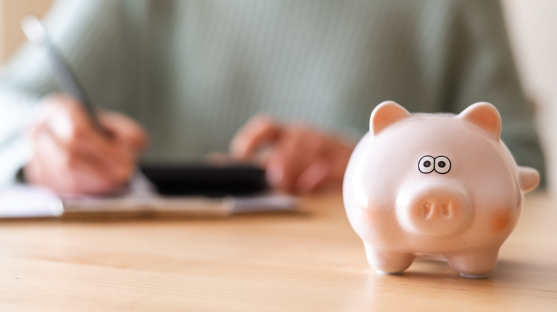 Woman calculating expenses with a calculator and notepad, with piggy bank on the table.