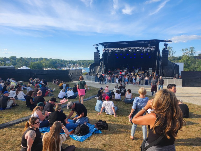 An outdoor stage on a summer day. A bay is visible in the background, and people are hanging around in casual clothes, waiting for a show to start.