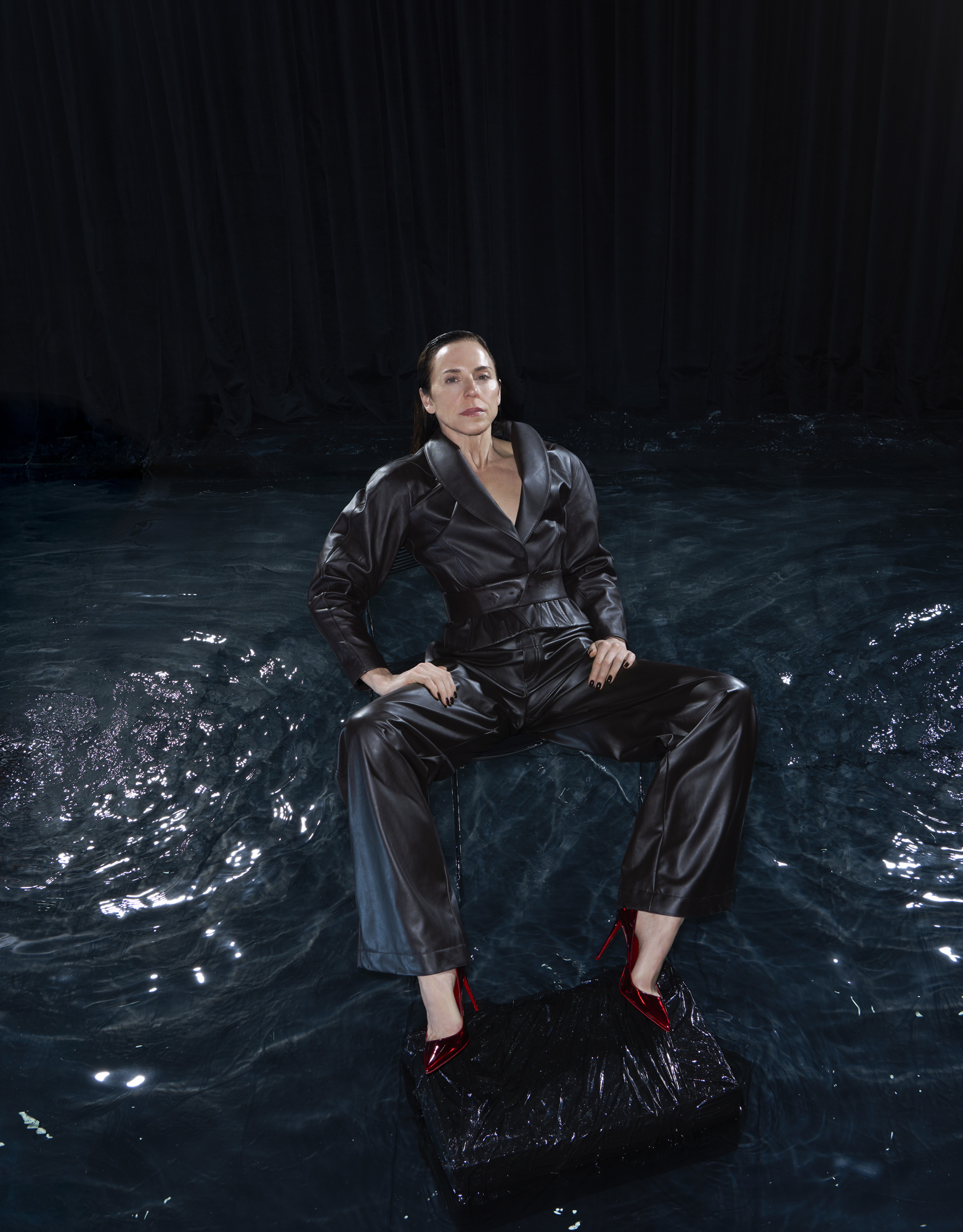 Melanie Chisholm in a dark leather pantsuit and red heels, sitting in dark water.