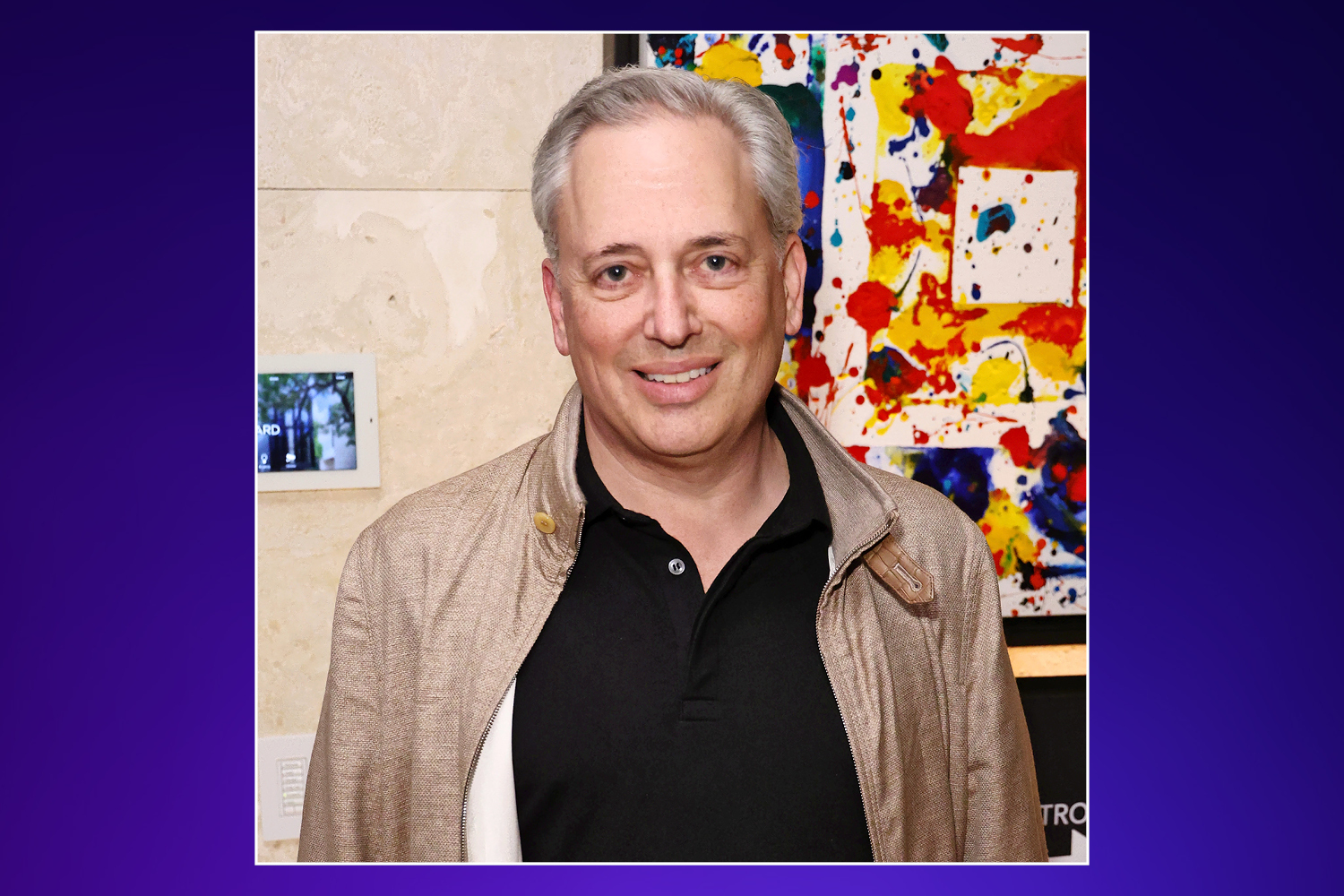 A man with gray hair and a beige jacket over a black shirt smiles at the camera, with abstract art behind him.