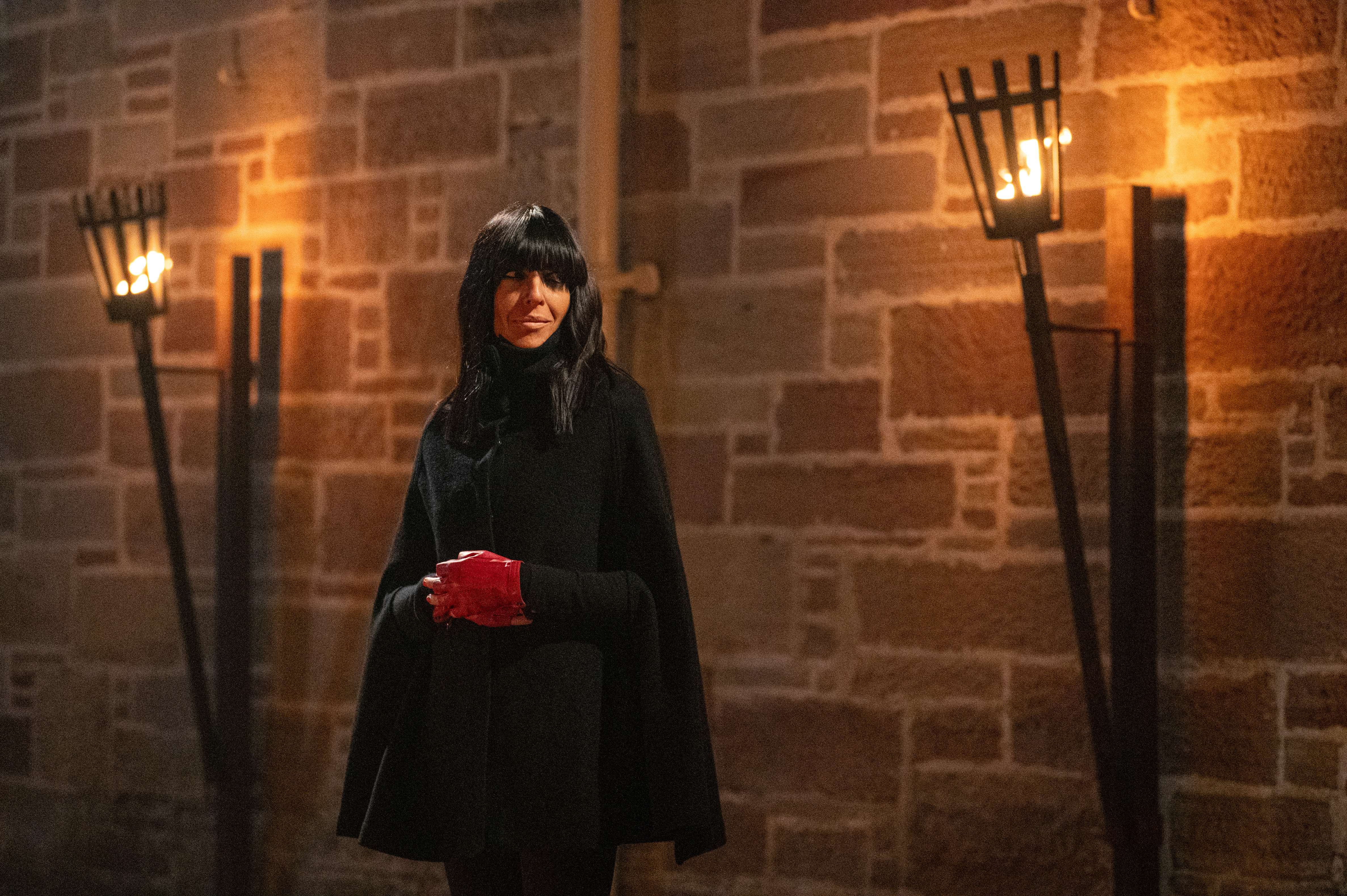 Claudia Winkleman in a black cape and red gloves, standing in front of a stone wall illuminated by two torches.