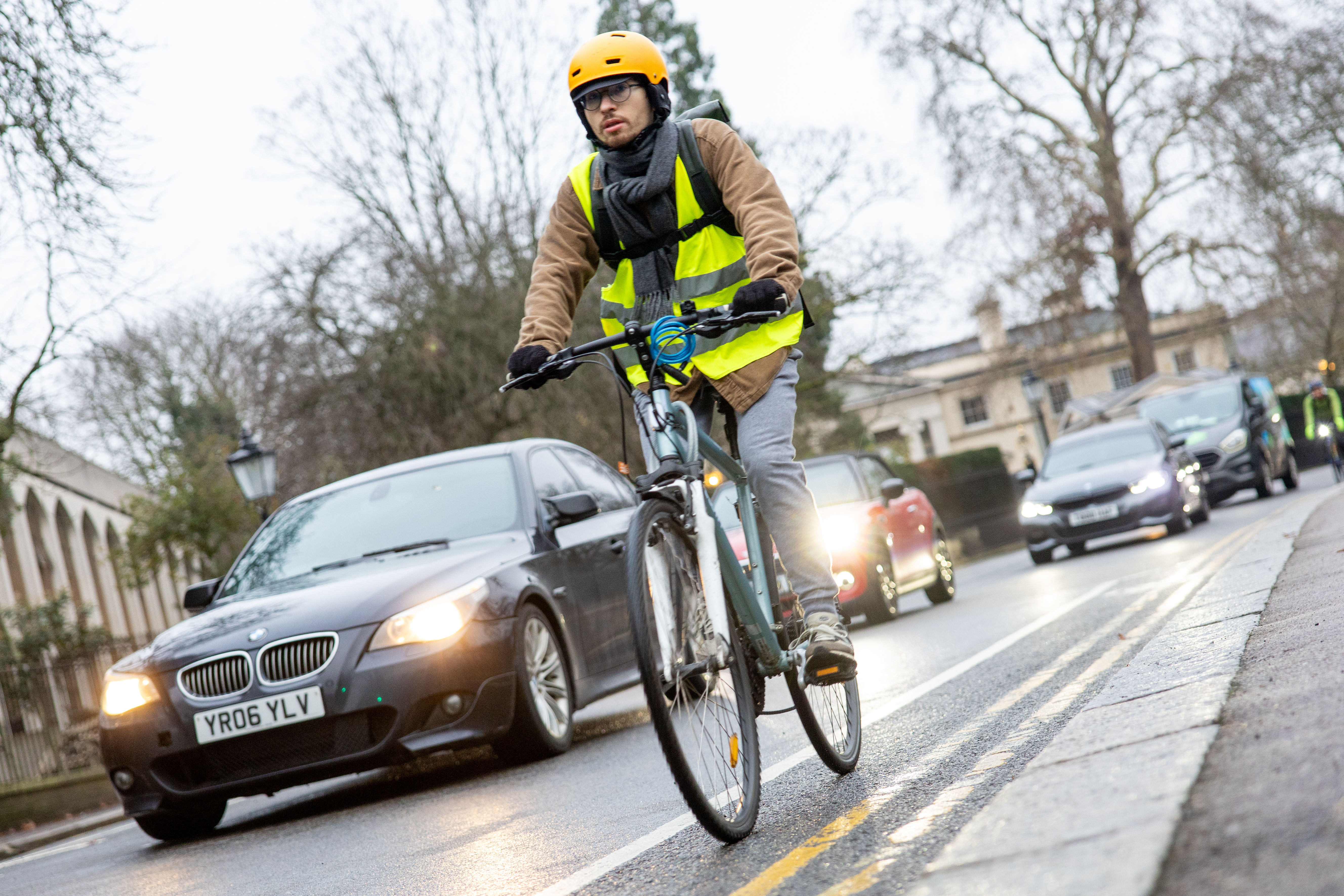 A cyclist in a yellow helmet and safety vest rides alongside cars in Regents Park.